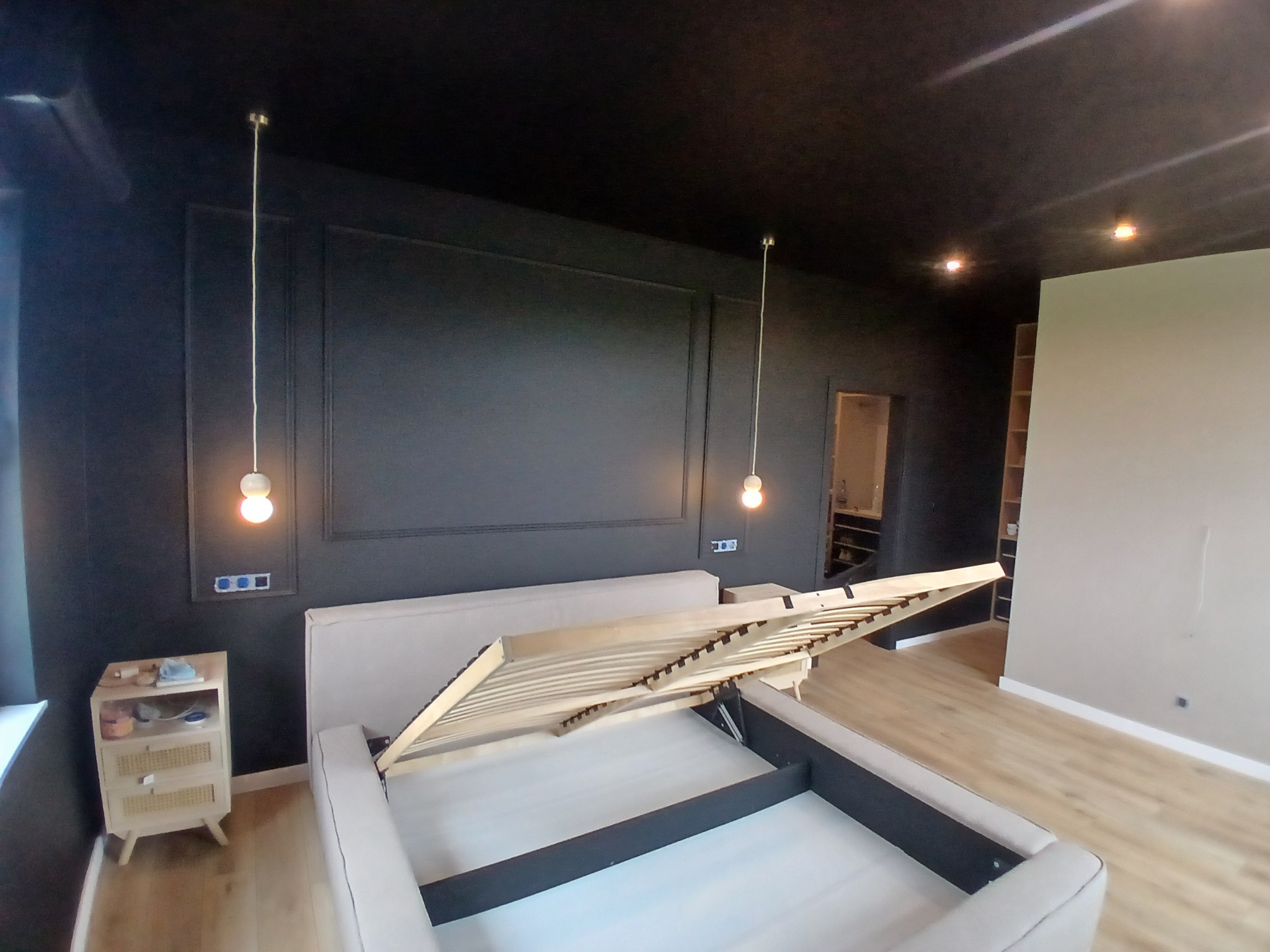 Elegancka sypialnia z czarnymi ścianami i sufitem, beżowe łóżko z podnoszonym stelażem, drewniana podłoga, minimalistyczne lampy wiszące i szafka nocna.