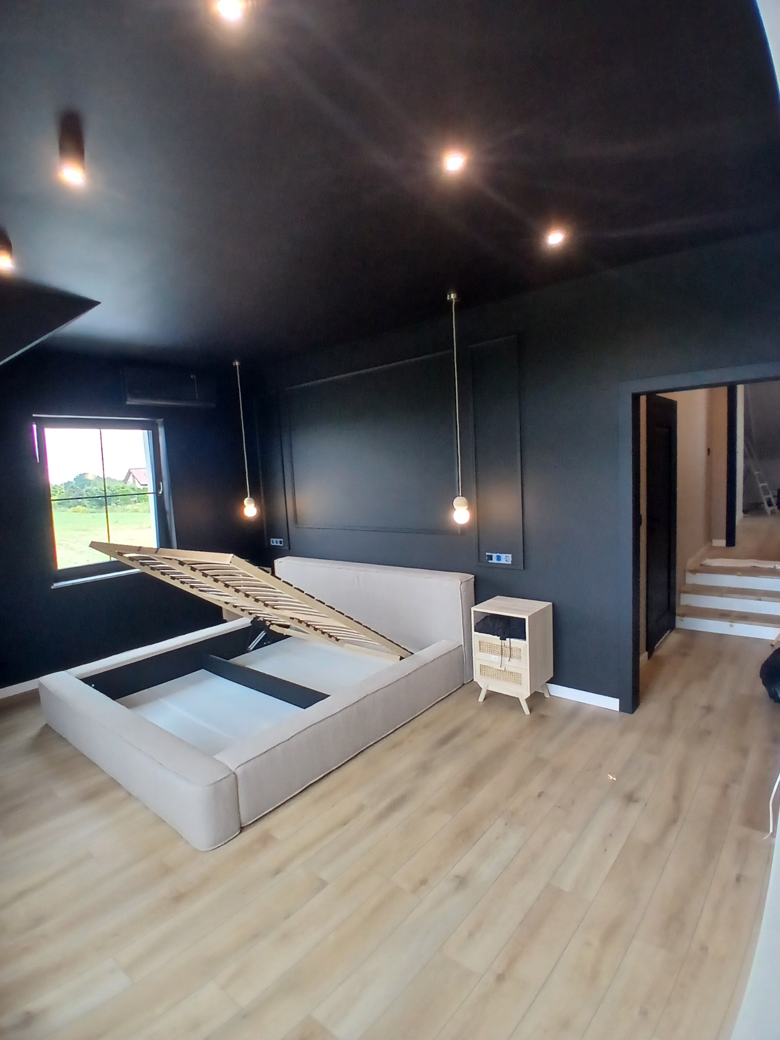 Ciemne wnętrze sypialni z beżowym łóżkiem z podnoszonym stelażem, drewnianą podłogą i wiszącymi lampami. Widok na okno i schody w tle.