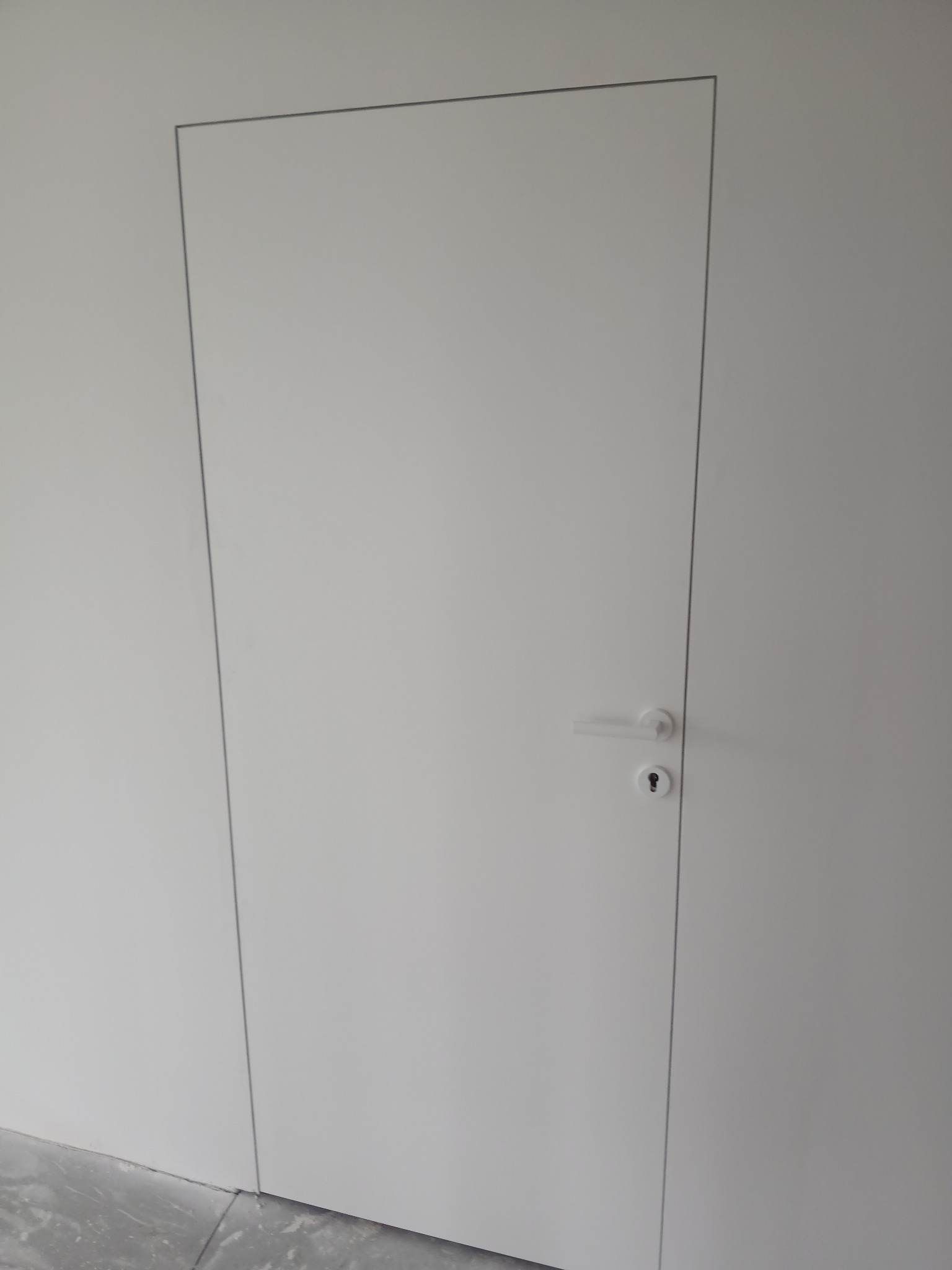 Minimalistyczne, białe drzwi ukryte w ścianie, z delikatną ramką i prostą klamką, tworzące jednolitą powierzchnię. Widoczny fragment podłogi.