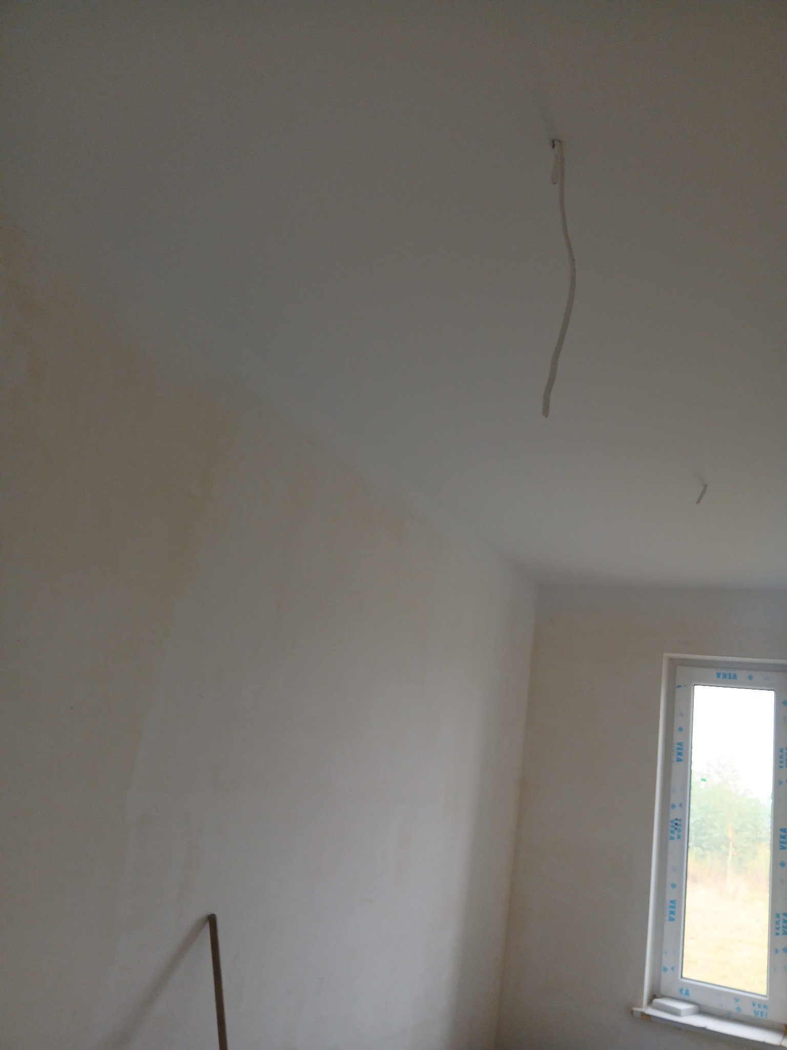 Ściana po nałożeniu gładzi, widoczne niedoskonałości i kabel zwisający z sufitu. Okno z widokiem na zewnątrz. Przy ścianie oparty kij.