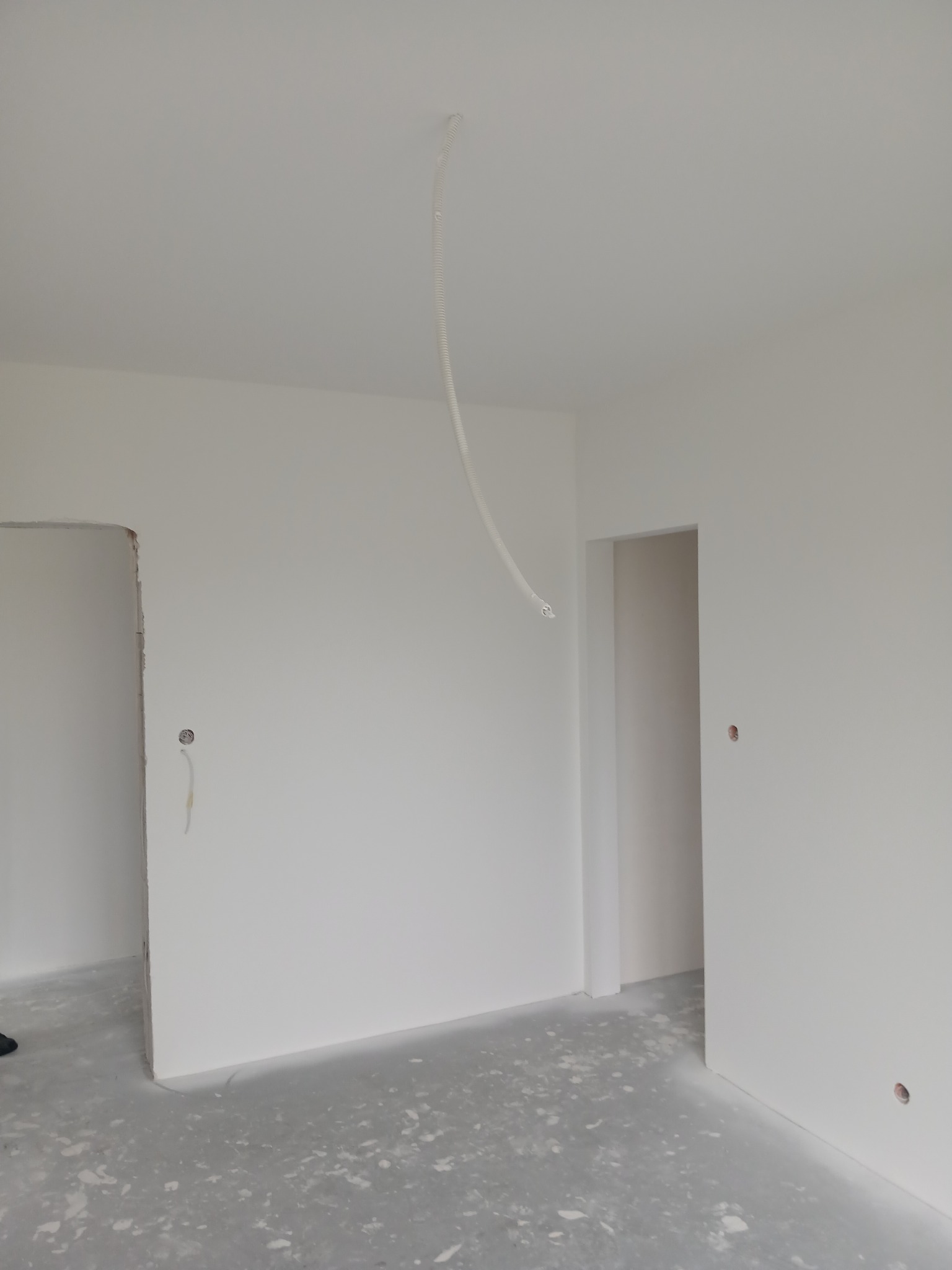 Wnętrze w trakcie prac wykończeniowych: białe ściany, surowa podłoga, widoczne otwory na gniazdka i peszel zwisający z sufitu. Minimalistyczny kadr, surowe wykończenie.