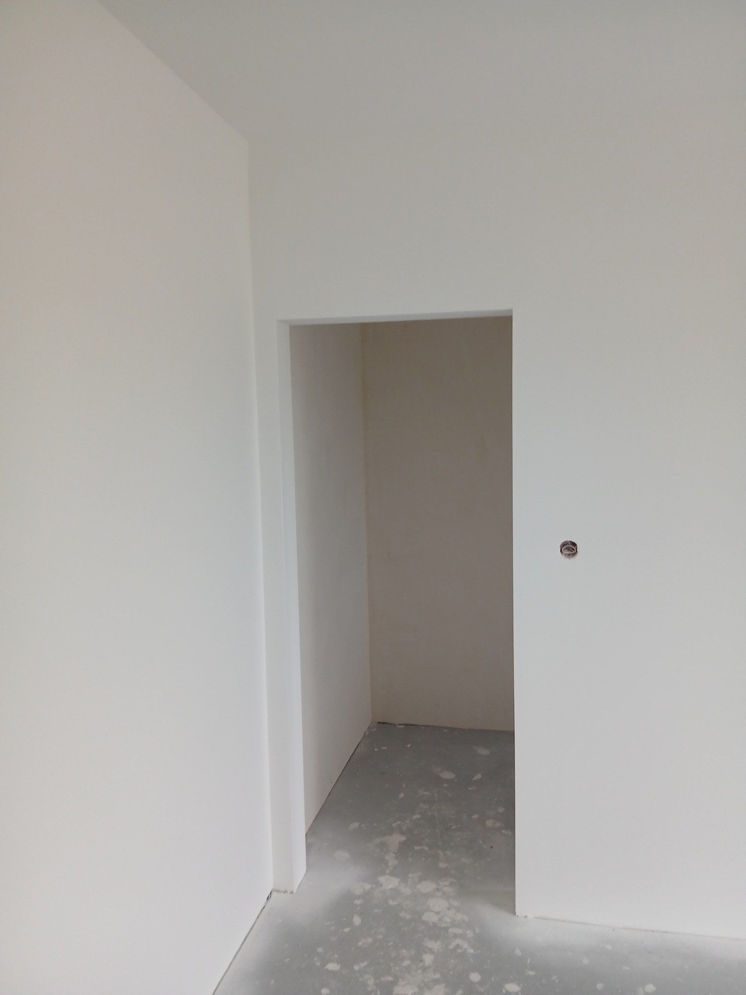 Białe, świeżo pomalowane ściany z widocznym otworem drzwiowym i szarym betonowym podłożem. Minimalistyczne wykończenie wnętrza, surowy stan po malowaniu.