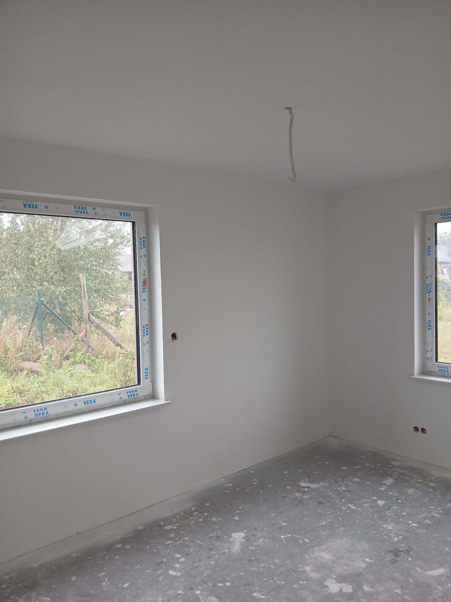 Wnętrze po szpachlowaniu: gładkie, białe ściany, okna PCV z widokiem na zieleń, surowa podłoga. Przygotowane do dalszych prac wykończeniowych. Widoczny kabel wystający z sufitu.