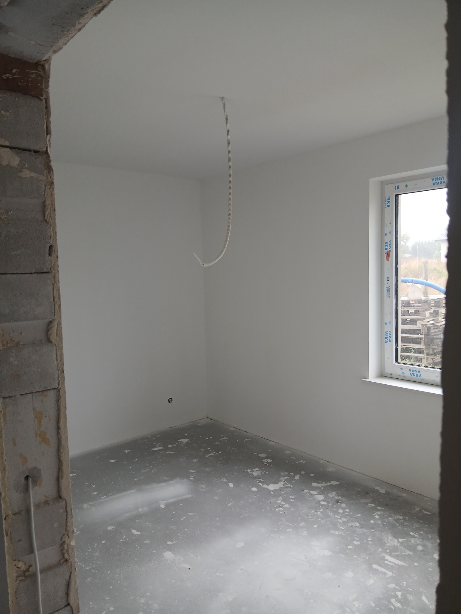 Wnętrze pokoju w trakcie prac wykończeniowych: świeżo pomalowane ściany na biało, surowa podłoga, okno z widokiem na zewnątrz i wystający kabel z sufitu.