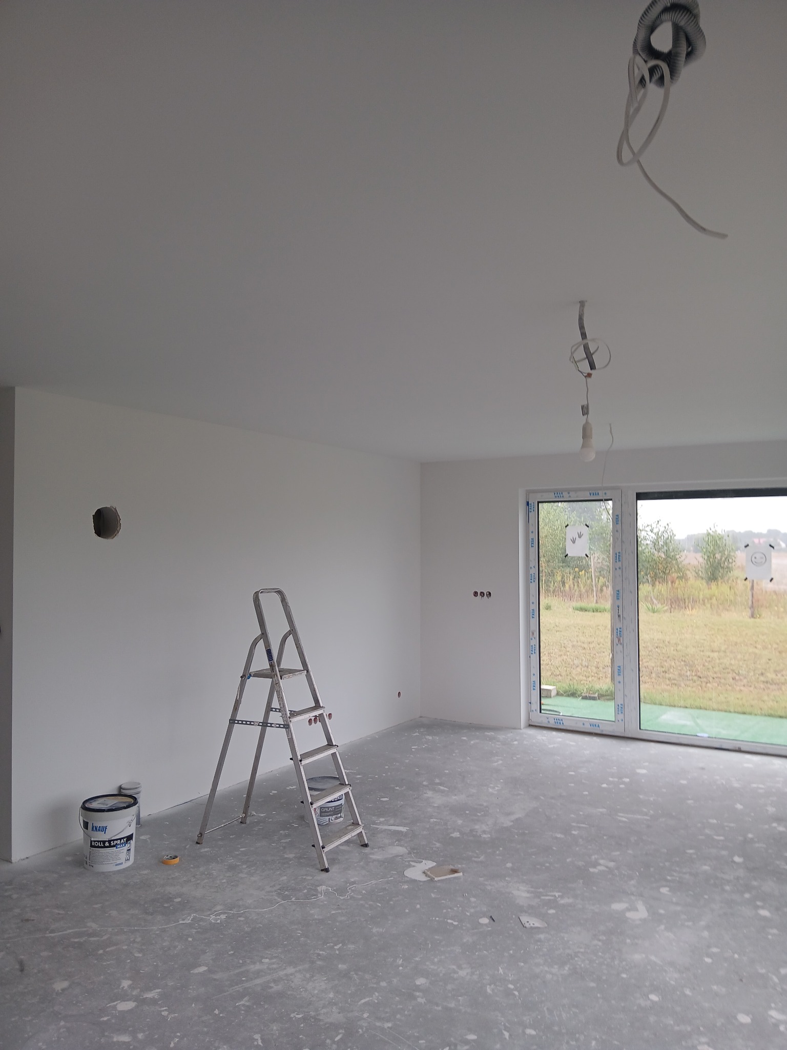 Wnętrze w trakcie malowania w Świeciu. Widoczna drabina, puszka z farbą i surowe wykończenie podłogi. Ściany świeżo pomalowane na biało, okno z widokiem na zewnątrz.