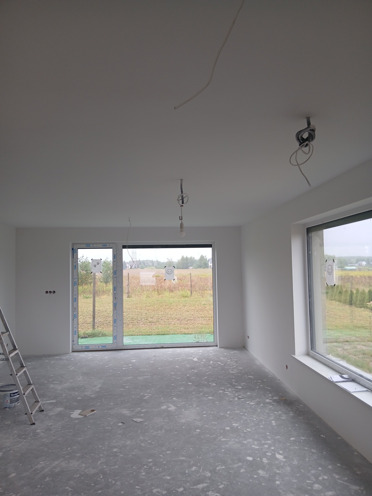 Jasne wnętrze w trakcie prac wykończeniowych: białe ściany, surowa podłoga, drabina oparta o ścianę, okna z widokiem na zewnątrz. Widoczne kable oświetleniowe.