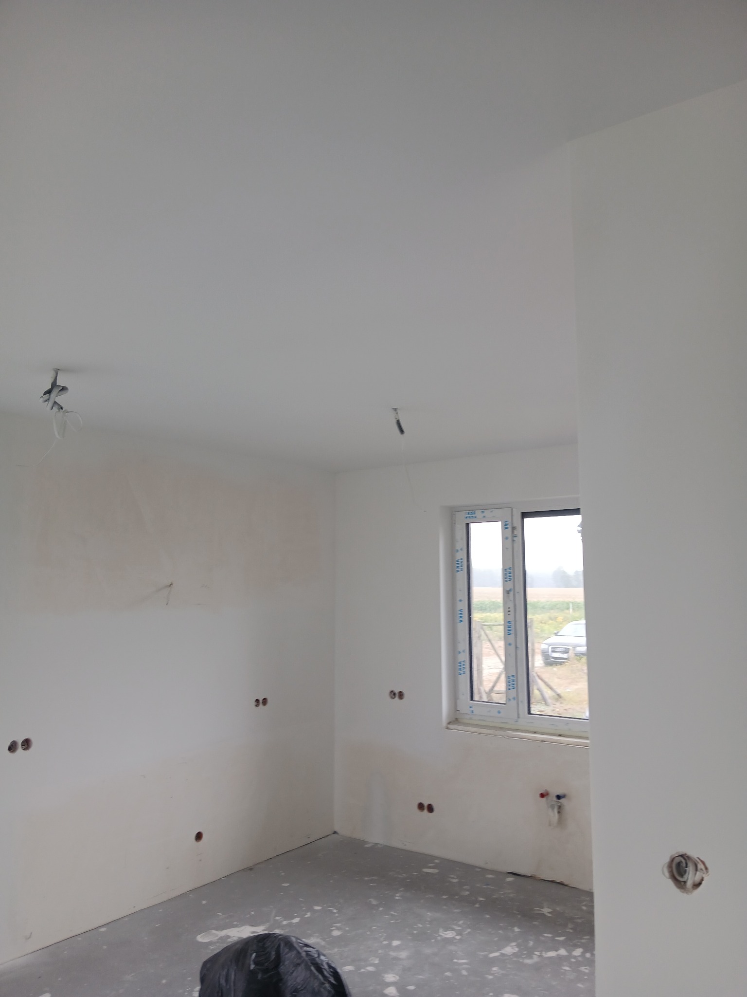 Wnętrze w trakcie prac: ściany częściowo pokryte gładzią, widoczne otwory na gniazdka, okno z widokiem na zewnątrz. Podłoga surowa, zabezpieczona folią.
