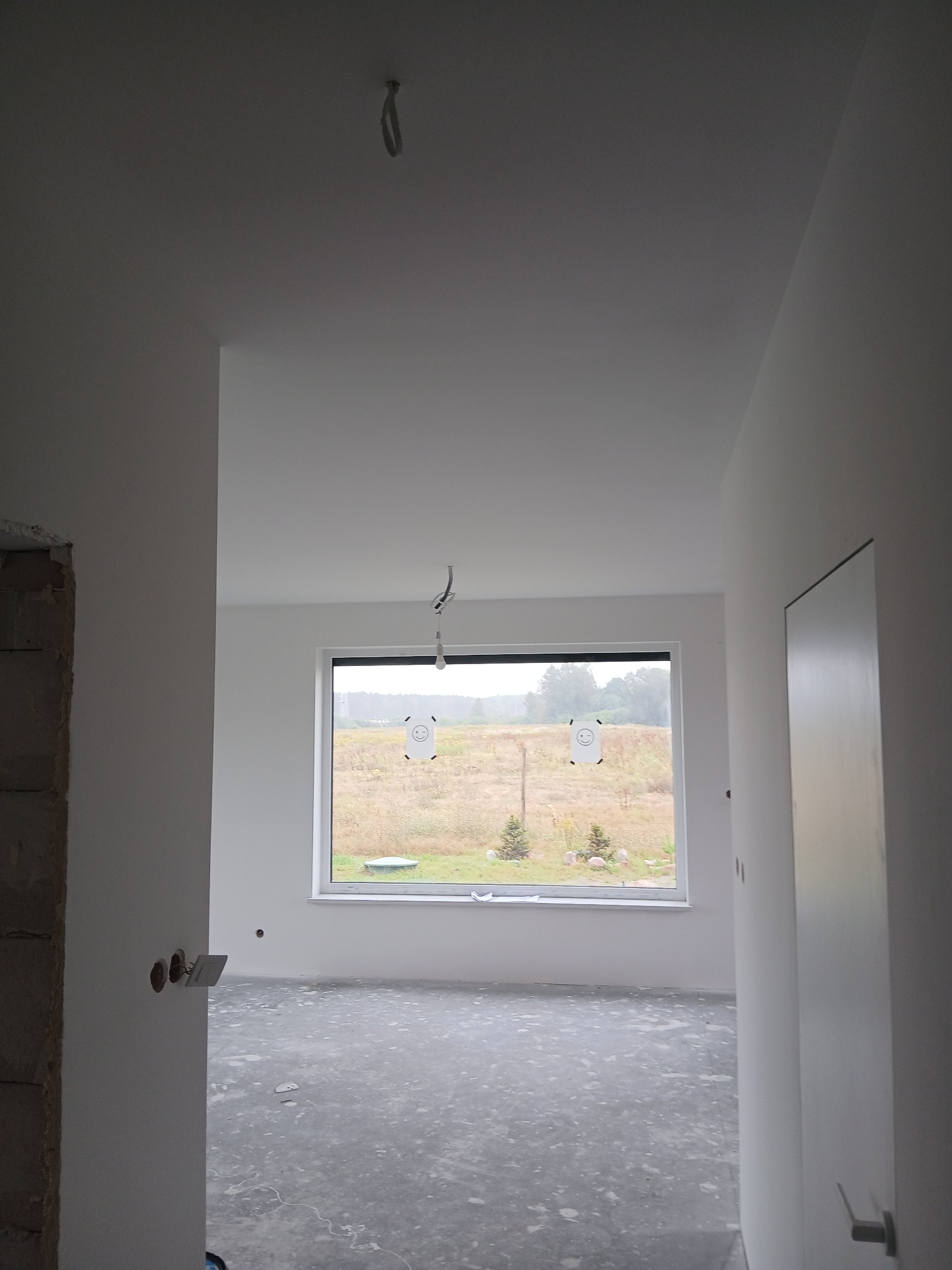 Wnętrze w stanie deweloperskim z oknem na zewnątrz. Widoczne ściany po gładziowaniu, sufit z wystającymi kablami i surowa podłoga. W tle widok na zewnątrz.