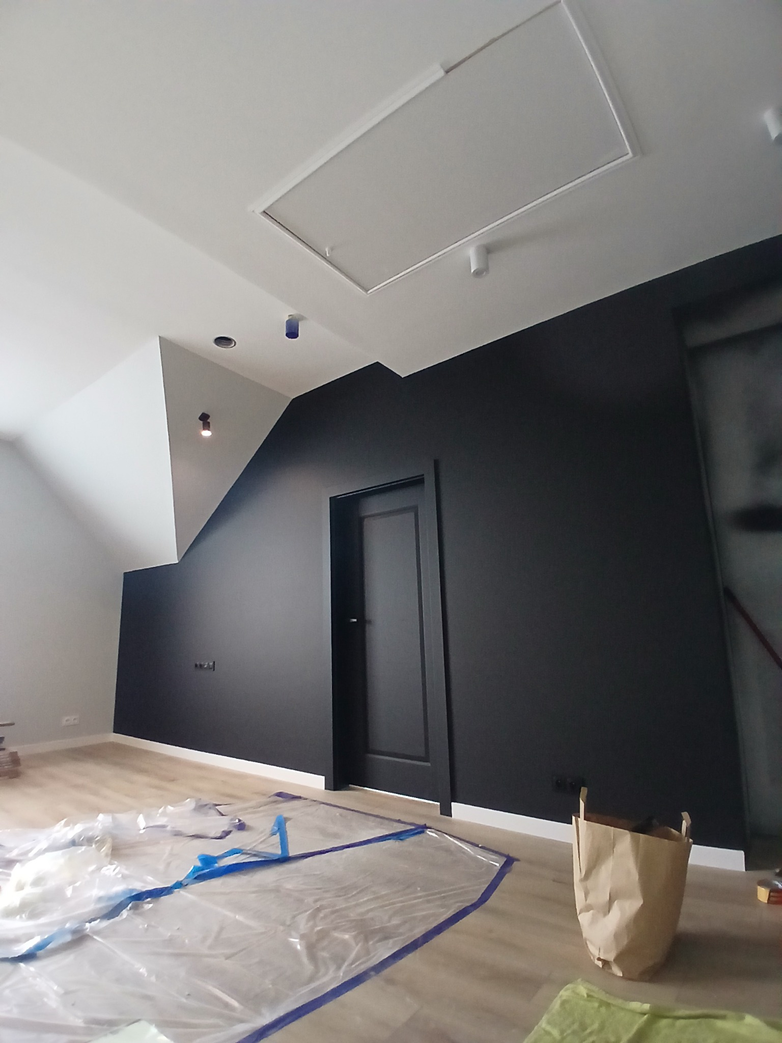 Wykończone wnętrze poddasza: czarna ściana z drzwiami, biały sufit z klapą, zabezpieczona podłoga. Minimalistyczny design, po malowaniu, widoczne detale wykończeniowe.
