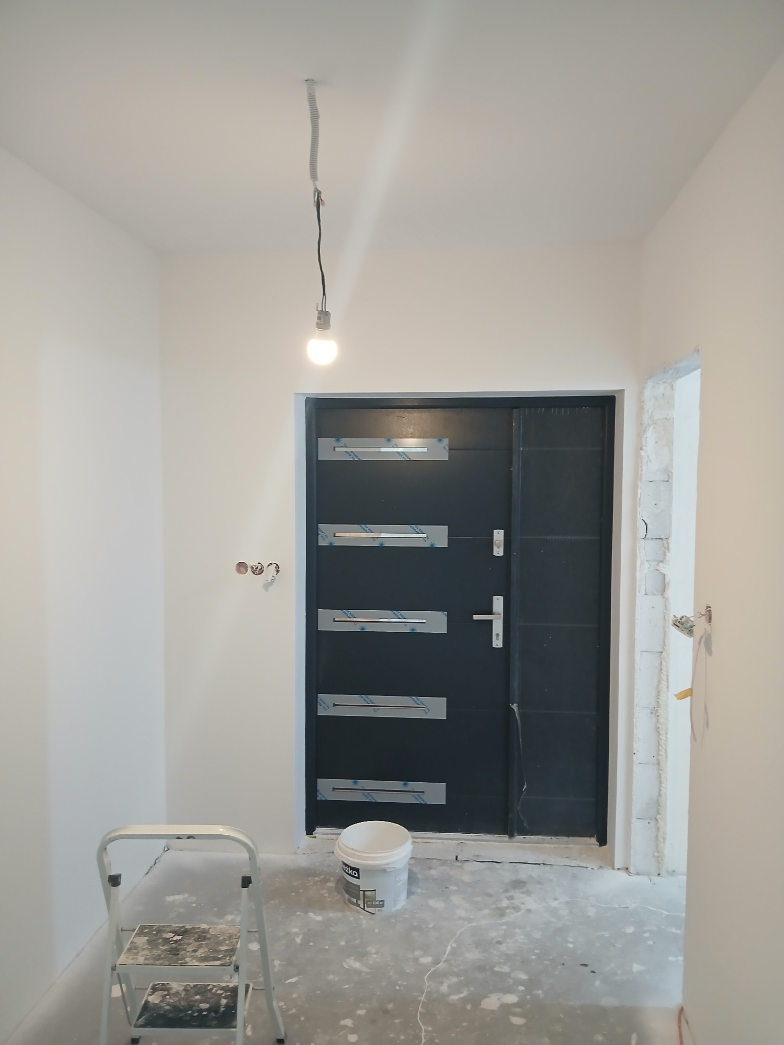 Wnętrze w trakcie odświeżania: białe ściany, czarne drzwi z folią ochronną, drabina malarska i wiadro farby na podłodze. Widoczne gniazdka elektryczne i surowy otwór drzwiowy.