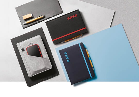 Stylowe kalendarze firmowe w różnych kolorach i wzorach, leżące na kontrastowym tle, z dodatkami jak długopisy i telefon w kieszeni kalendarza.