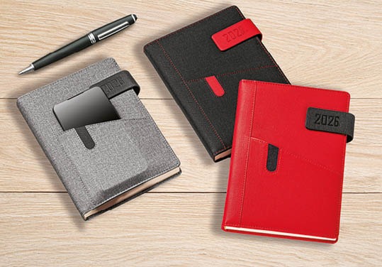 Trzy eleganckie kalendarze na rok 2026 w kolorach szarym, czarnym i czerwonym, leżące na drewnianym stole obok czarnego długopisu.