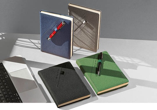 Cztery teksturowane kalendarze w odcieniach granatu, szarości, czerni i zieleni, każdy z dopasowanym długopisem, leżą na jasnym tle obok laptopa.