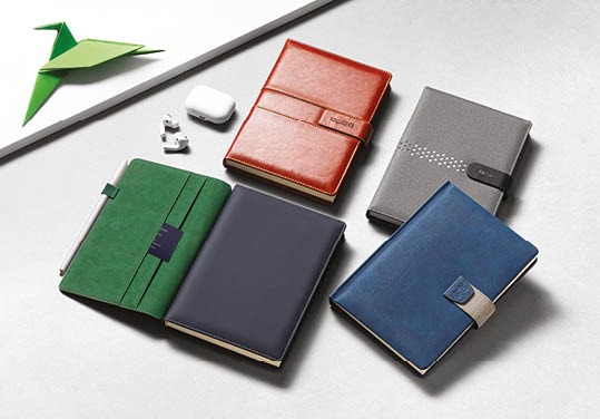 Eleganckie kalendarze z ekoskóry w różnych kolorach leżące na jasnym tle, obok słuchawki i origami. Widok z góry.