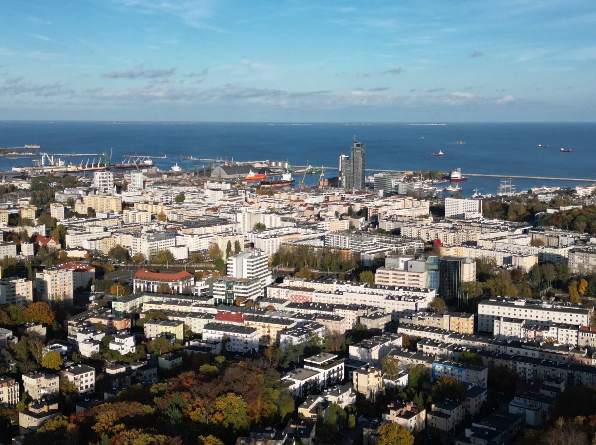 Panorama Gdyni z widokiem na morze, port i Sea Towers. Jesienne drzewa w pierwszym planie, błękitne niebo z delikatnymi chmurami w tle. Ujęcie z lotu ptaka.