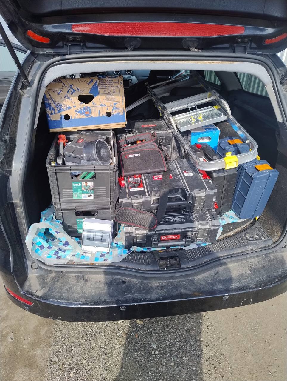 Otwarty bagażnik czarnego kombi wypełniony skrzynkami narzędziowymi, drabiną i rozdzielnicą elektryczną, sugerujący przygotowanie do pracy instalatora.