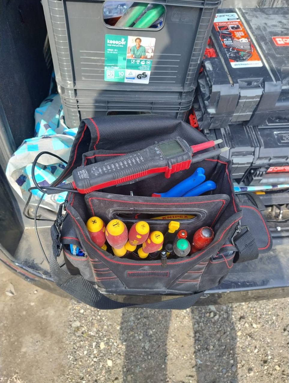 Torba narzędziowa Knipex z multimetrem, śrubokrętami i szczypcami na tle skrzynek narzędziowych i bagażnika samochodu, sugerująca gotowość do pracy.