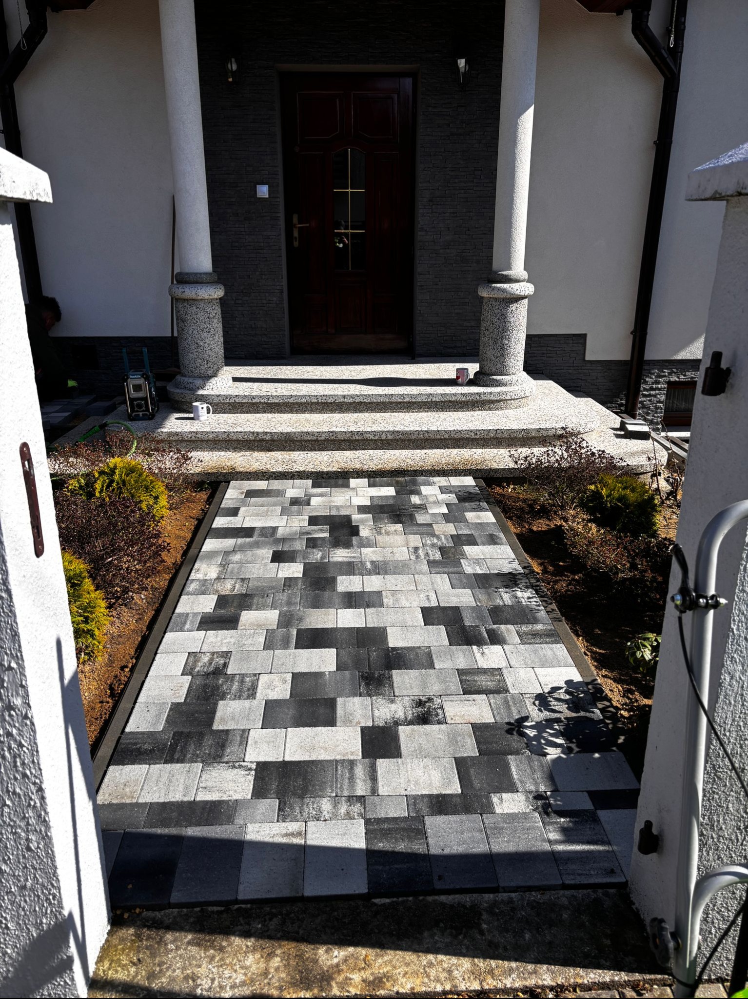 Ułożona ścieżka z kostki brukowej w odcieniach szarości prowadząca do wejścia domu z kolumnami. Widoczne obrzeża, roślinność i narzędzia pracy.