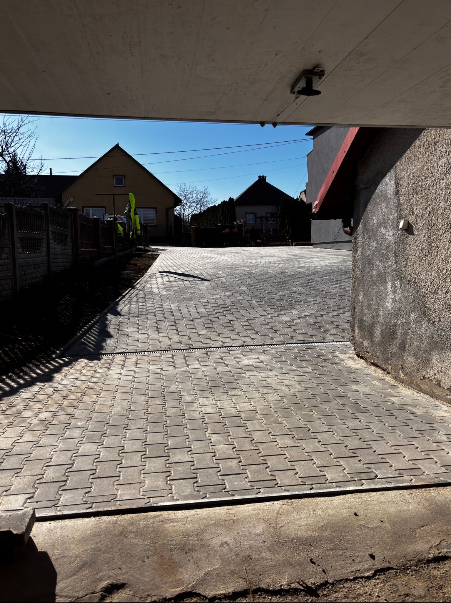 Szara kostka brukowa ułożona na podjeździe, widok z zadaszonego wjazdu. W tle domy jednorodzinne i błękitne niebo. Widoczny drenaż liniowy.