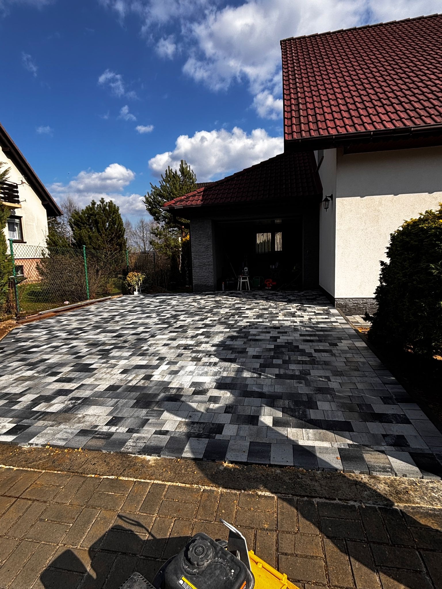 Ułożona kostka brukowa w szaro-białej mozaice przed domem z czerwonym dachem. W tle widoczne drzewa i błękitne niebo z chmurami. Na pierwszym planie fragment maszyny.