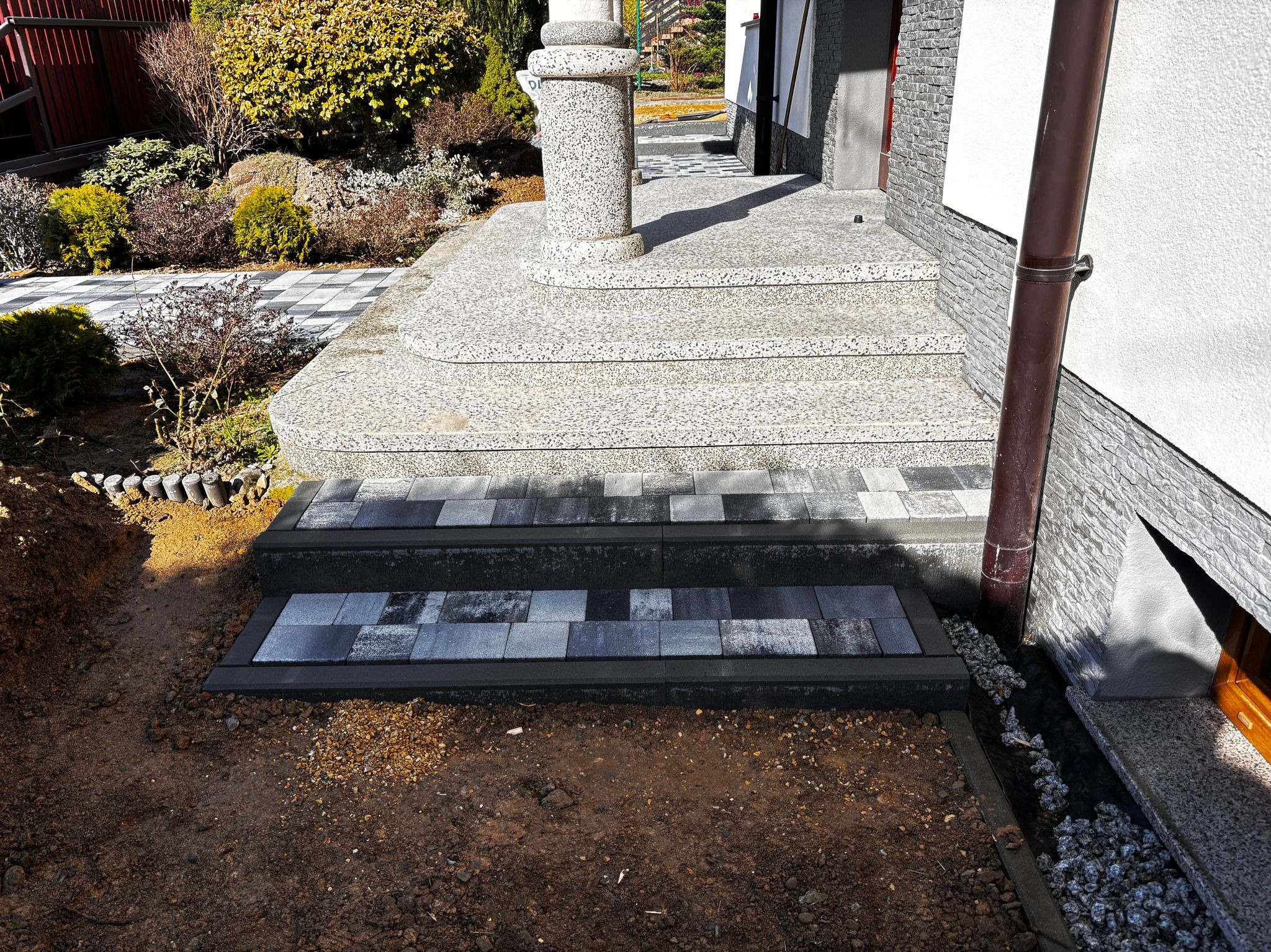 Schody wejściowe z kostki brukowej w odcieniach szarości, prowadzące do domu z kolumną. Częściowo wykopana ziemia wokół schodów.