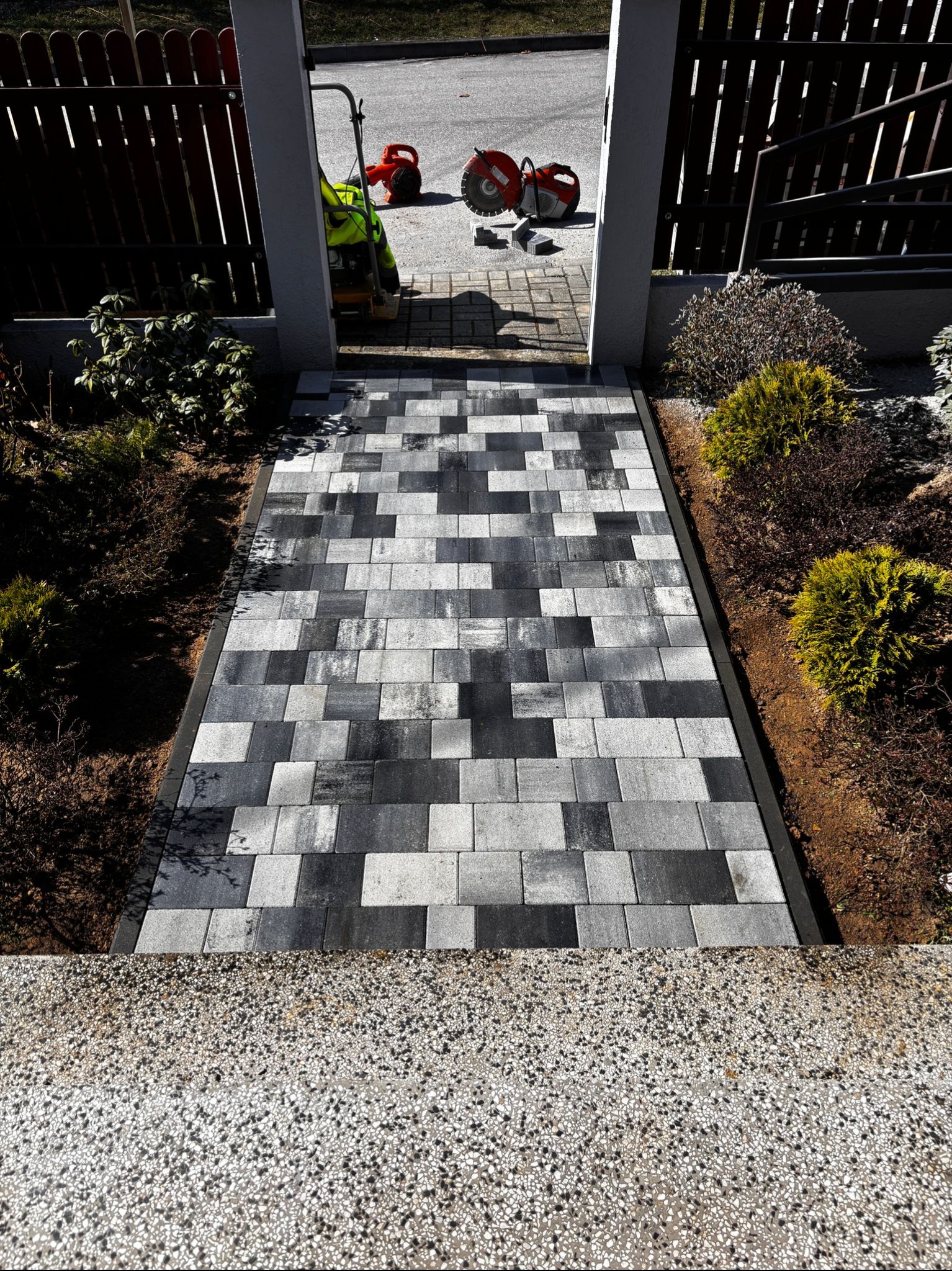 Ułożona ścieżka z kostki brukowej w odcieniach szarości, prowadząca przez bramkę. W tle widoczne narzędzia i maszyny budowlane, sugerujące etap końcowy prac.
