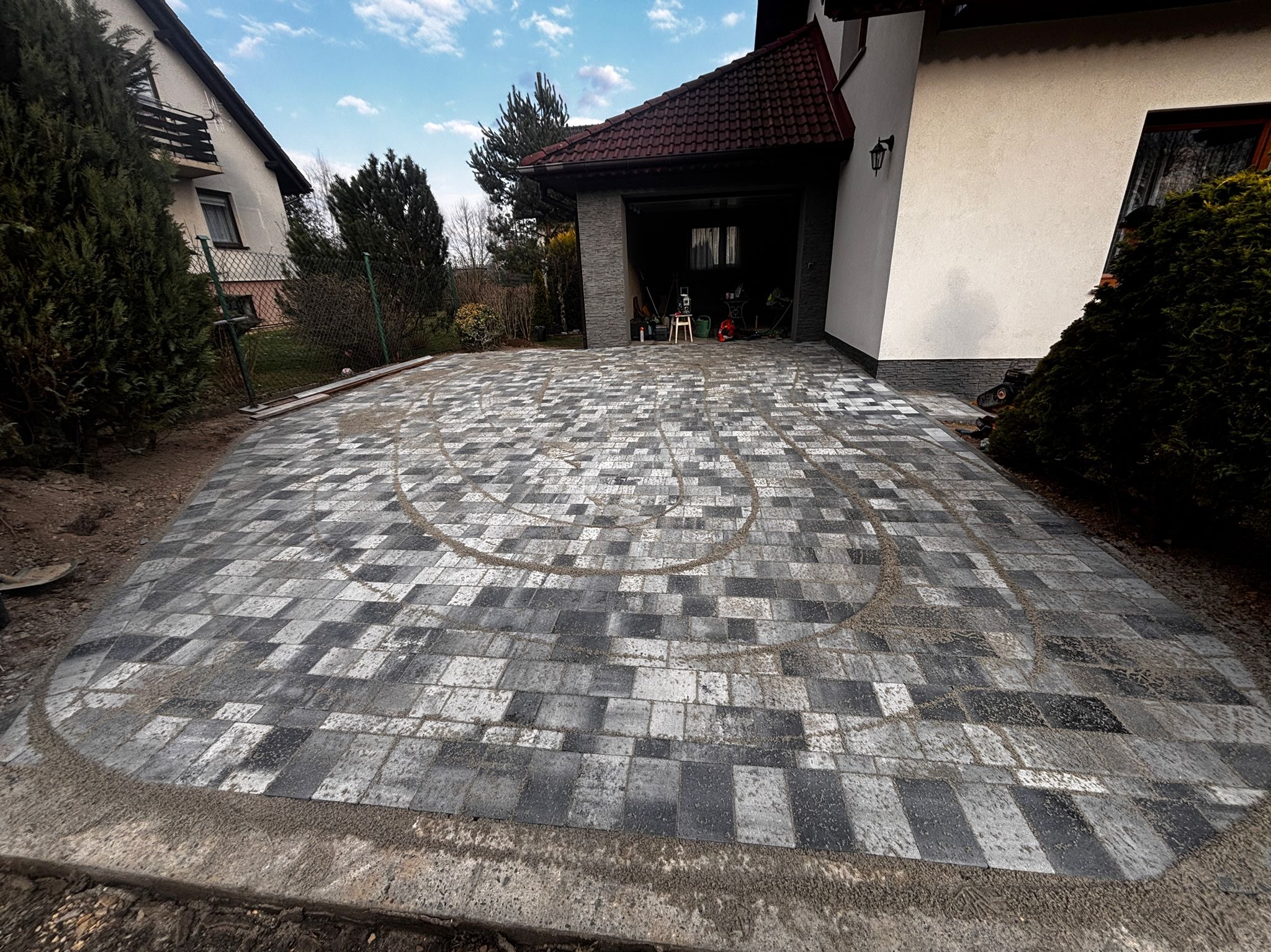 Ułożona kostka brukowa w odcieniach szarości tworząca wzór na podjeździe przed domem z otwartym garażem. Widoczne ślady piasku.