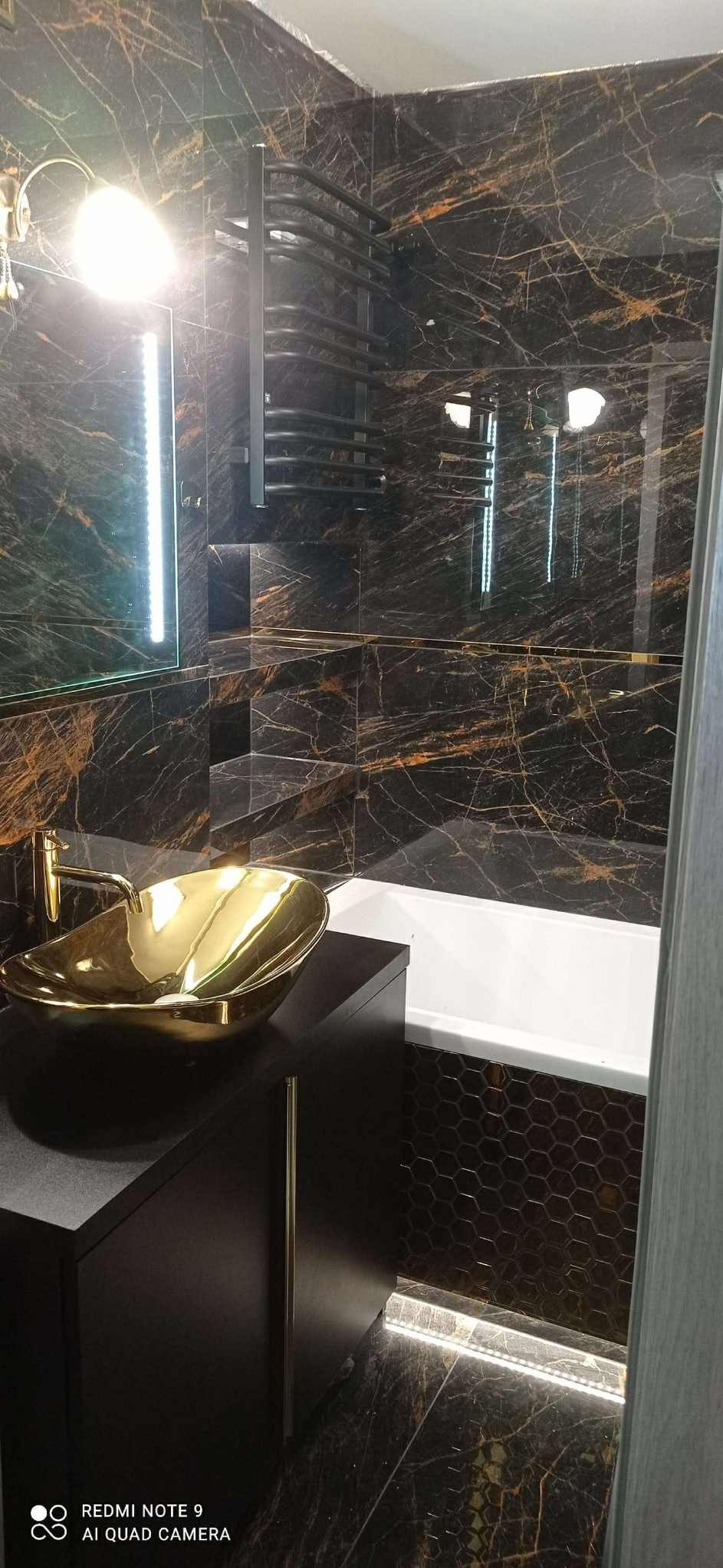Elegancka łazienka z czarnymi płytkami imitującymi marmur, złotą umywalką i grzejnikiem drabinkowym. Nowoczesne oświetlenie LED podkreśla detale.