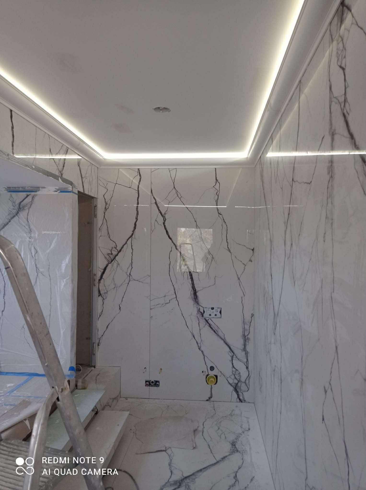 Wykończona łazienka z białymi płytkami imitującymi marmur, oświetleniem LED podsufitowym i aluminiową drabiną w rogu. Widoczne gniazdka i instalacje.