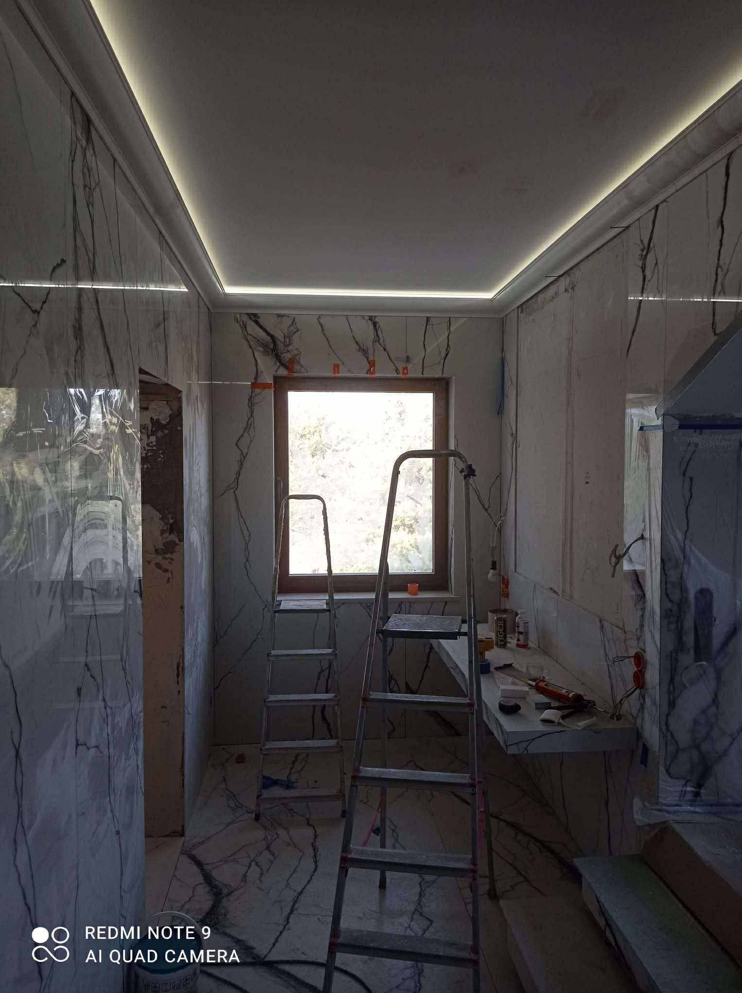Łazienka w trakcie wykończenia: marmurowe płytki na ścianach i podłodze, drabina, okno. Widoczne narzędzia i materiały budowlane. Oświetlenie LED na suficie.