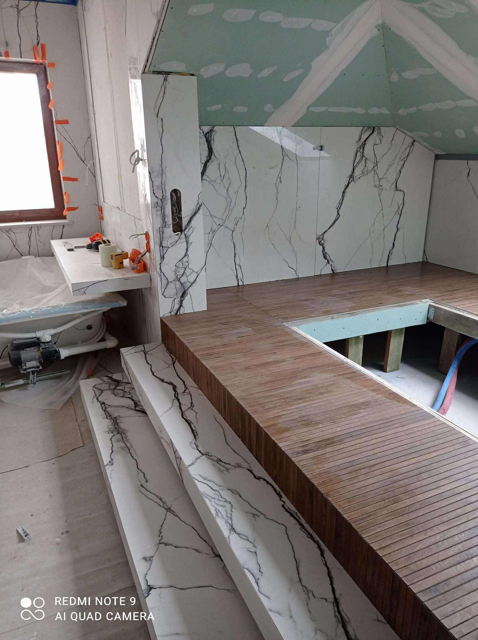 Łazienka w trakcie wykończenia: marmurowe płytki na ścianach, drewniana podłoga z desek, sufit z płyt kartonowo-gipsowych. Widoczne narzędzia i materiały budowlane.
