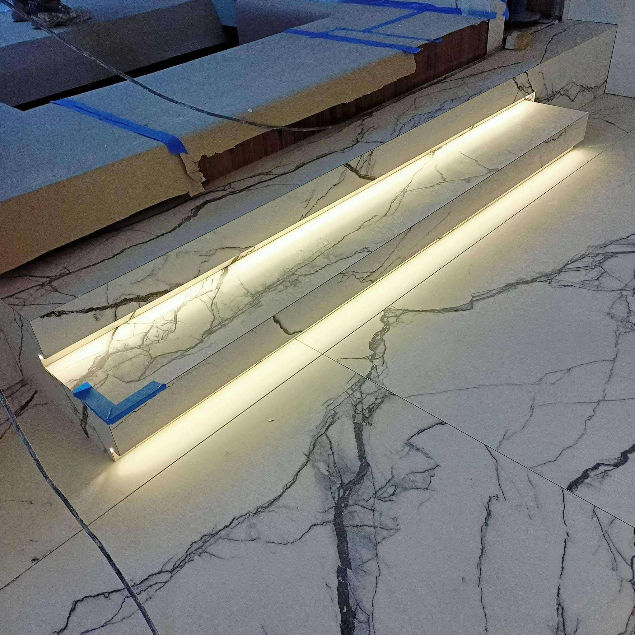 Podświetlane, marmurowe stopnie schodów w trakcie montażu, zabezpieczone niebieską taśmą. Widoczny fragment podłogi z płytek imitujących marmur.