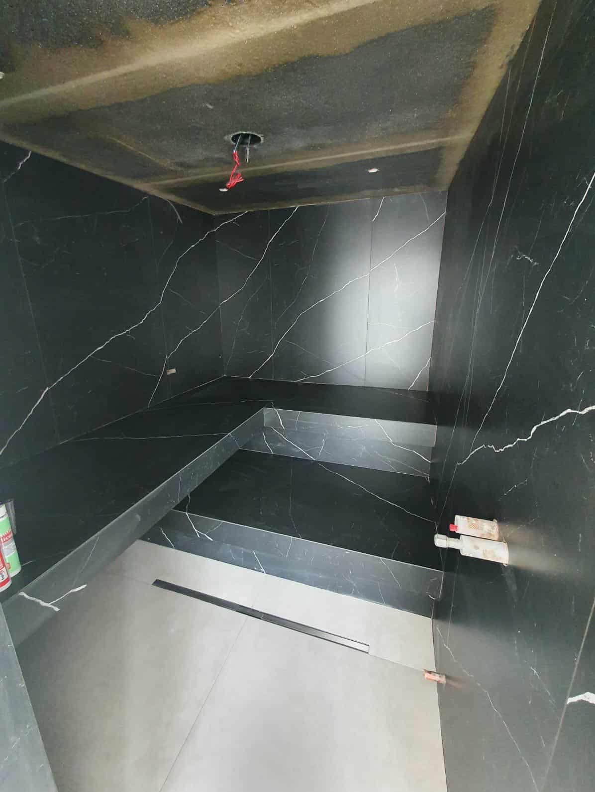 Nowoczesna łazienka z czarnymi płytkami imitującymi marmur, z wbudowanym siedziskiem i odpływem liniowym. Surowy beton na suficie, widoczne narzędzia glazurnicze.