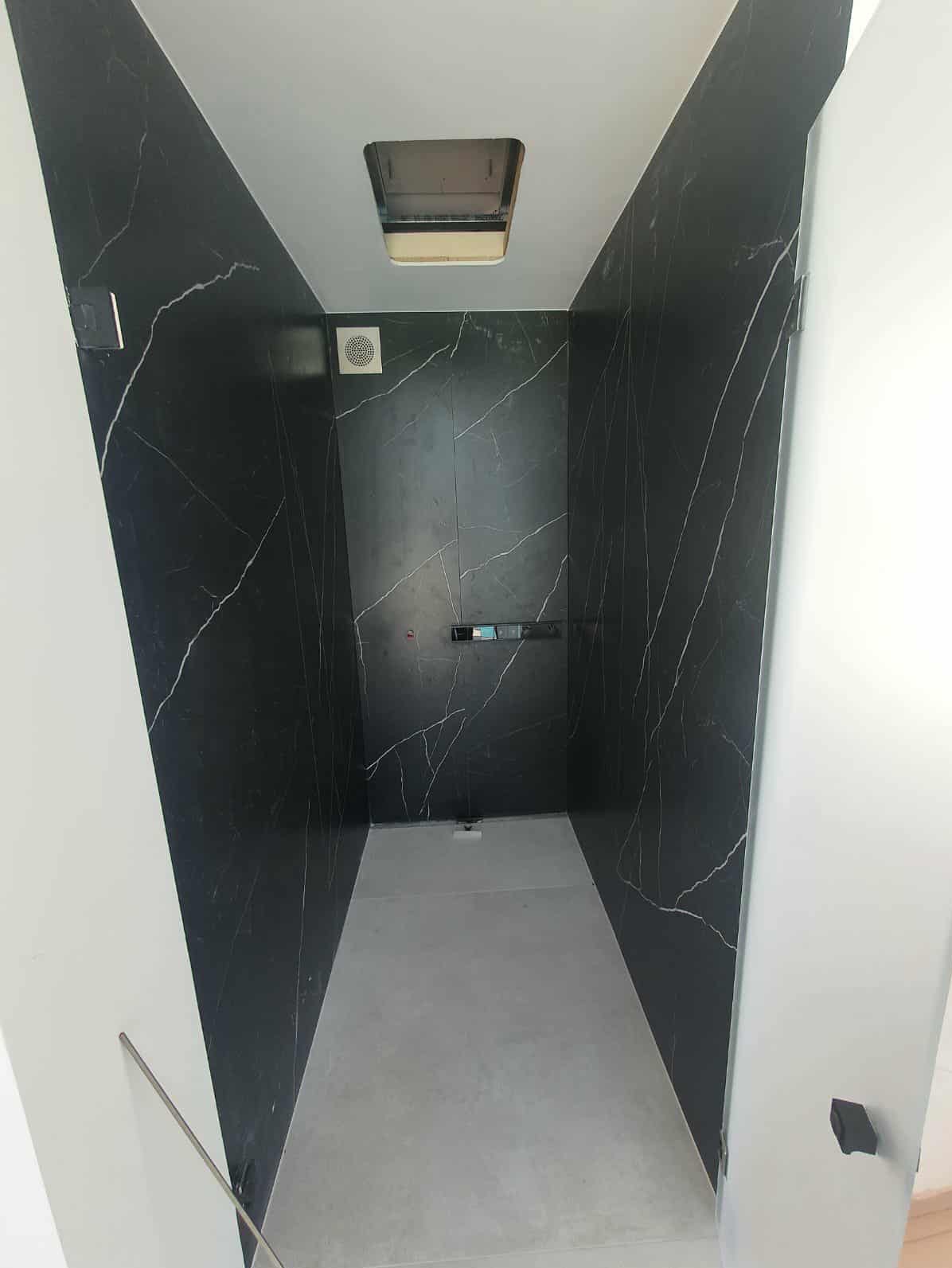 Nowoczesna kabina prysznicowa z czarnymi płytkami imitującymi marmur z białymi żyłami, szary brodzik, białe ściany i sufit z kratką wentylacyjną.