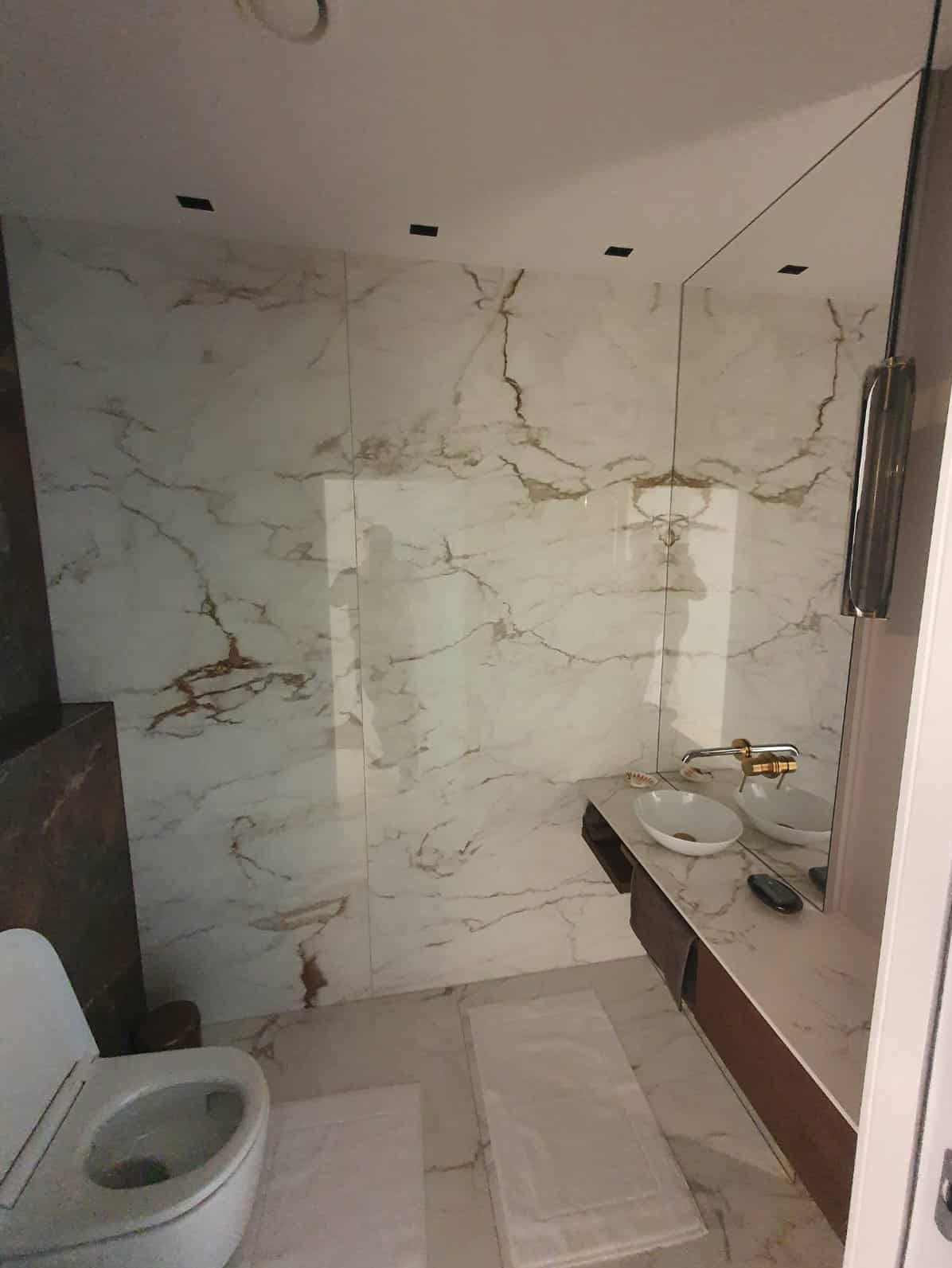 Elegancka łazienka z marmurowymi płytkami na ścianach i podłodze, umywalką nablatową i dużym lustrem. Widoczny fragment toalety i dywanik.