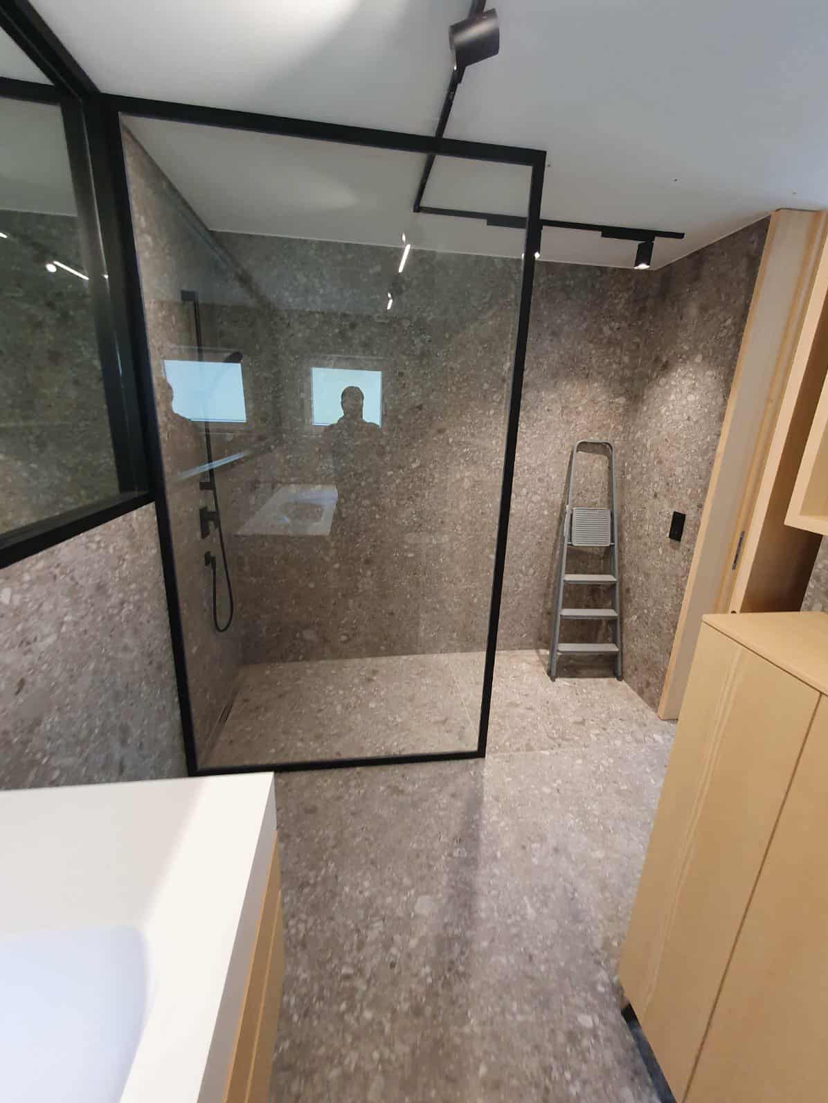 Nowoczesna kabina prysznicowa z czarną ramą i szklaną ścianą, wykończona płytkami imitującymi lastryko. Widoczna drabina i oświetlenie punktowe.