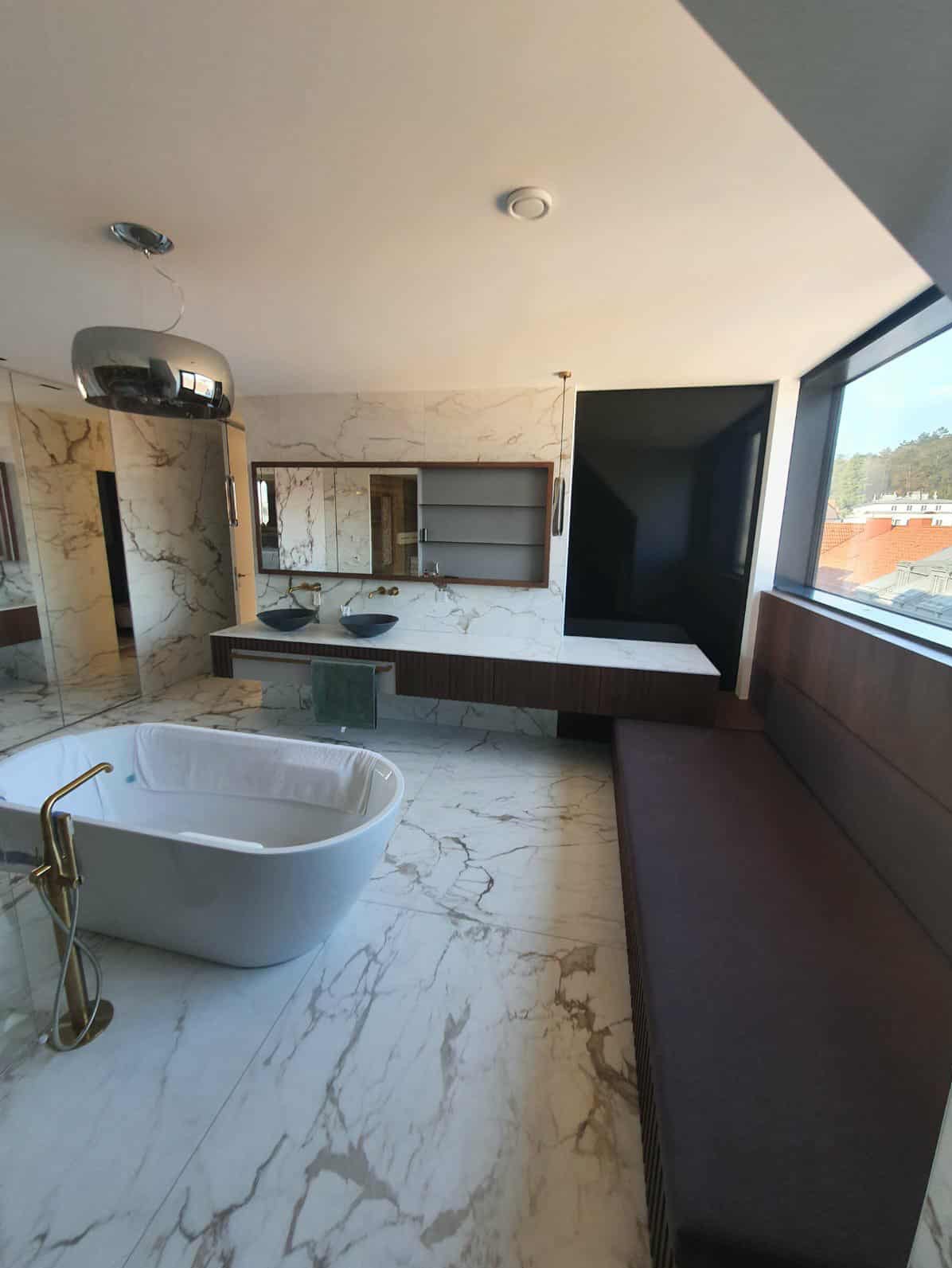 Elegancka łazienka z marmurową podłogą i ścianami, wolnostojąca wanna z złotą armaturą, dwie umywalki na blacie z szafką i duże okno z widokiem na zewnątrz.