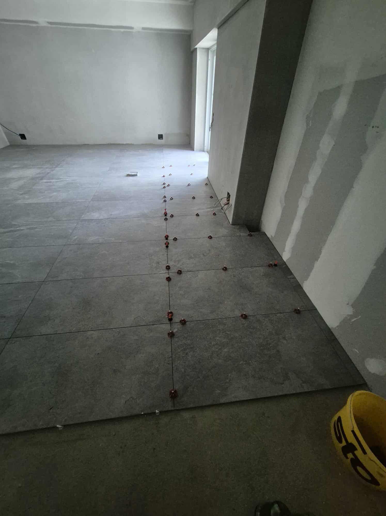 Układanie szarych płytek podłogowych w dużym formacie, z użyciem poziomujących krzyżyków. Ściany w surowym stanie, przygotowane do dalszych prac wykończeniowych.
