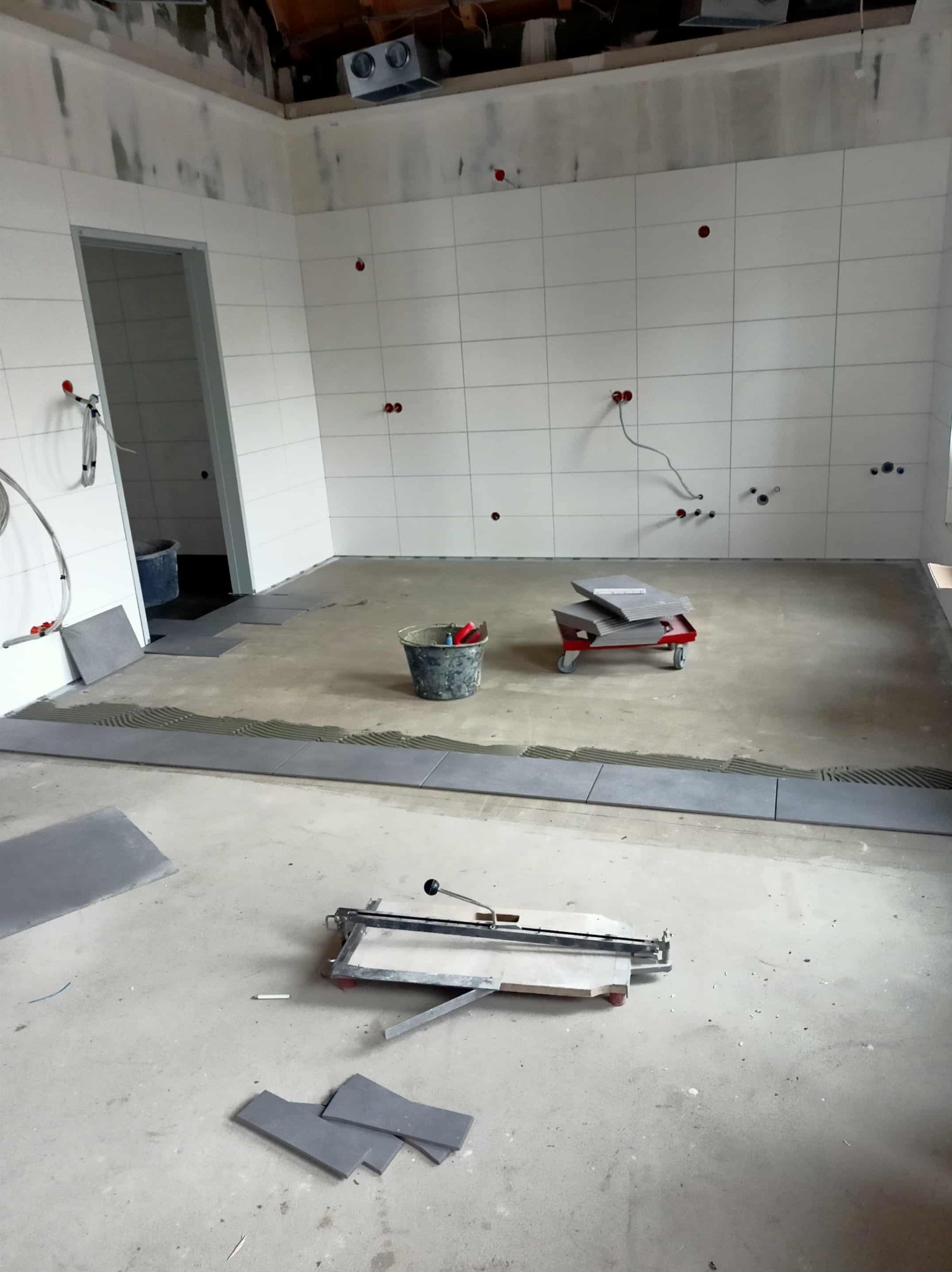 Wnętrze w trakcie układania płytek: białe płytki na ścianach, szare na podłodze. Widoczny sprzęt glazurniczy, wiadro z zaprawą i wózkiem z płytkami. Częściowo ułożone płytki podłogowe.