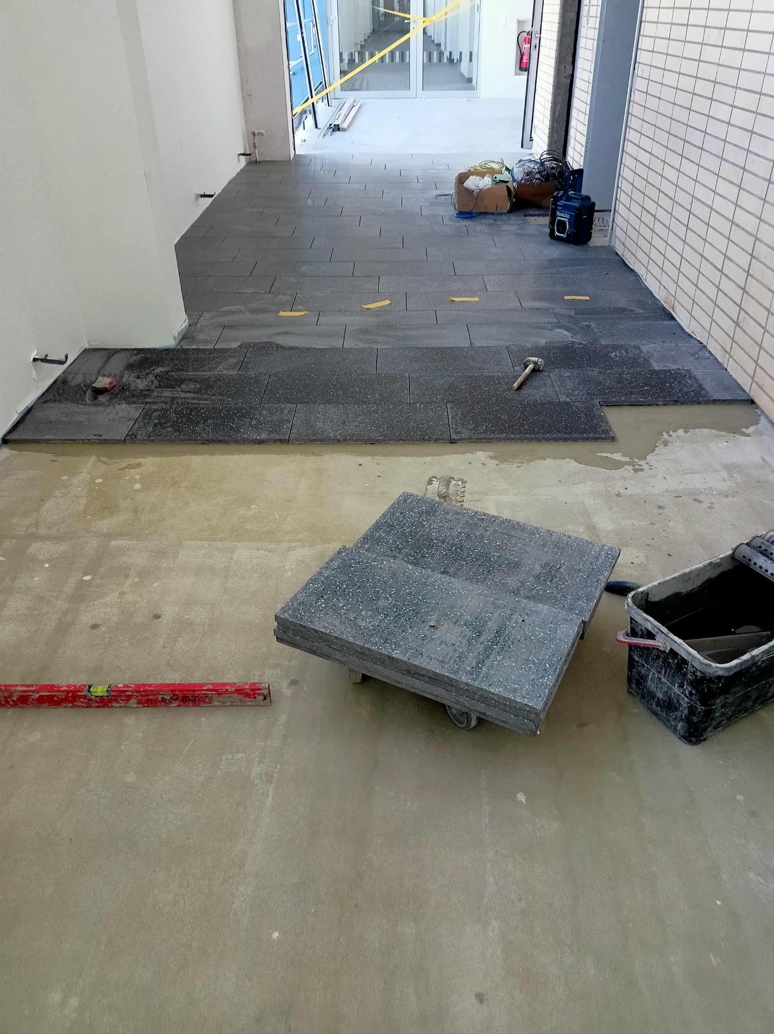 Układanie ciemnych płytek podłogowych w korytarzu w Kielcach. Widoczny poziomica, wiadro z zaprawą i stos płytek na wózku. Częściowo ułożona podłoga.