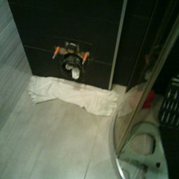 HydroHeros PAWEŁ PRASEK - Zamontowany stelaż podtynkowy WC z widocznymi przyłączami, zabezpieczony ręcznikiem, obok skrzynka z narzędziami. Ujęcie z góry, w łazience z szarymi płytkami.