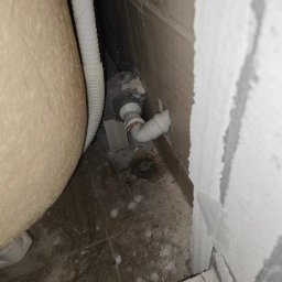 HydroHeros PAWEŁ PRASEK - Biała rura odpływowa podłączona do wanny, widoczna przy szarej ścianie z płytkami i surowym wykończeniem. Ujęcie z dołu, blisko podłogi.