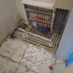 HydroHeros PAWEŁ PRASEK - Rozdzielacz ogrzewania podłogowego w szafce, z widocznymi rurami doprowadzającymi i odprowadzającymi wodę. Podłoga w trakcie prac wykończeniowych, widoczne ślady po kleju.