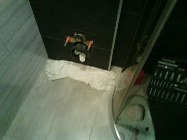 Zamontowany stelaż podtynkowy WC z widocznymi przyłączami, zabezpieczony ręcznikiem, obok skrzynka z narzędziami. Ujęcie z góry, w łazience z szarymi płytkami.