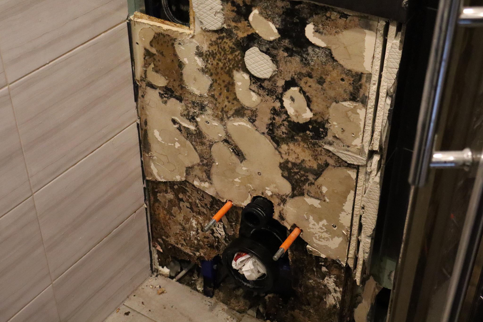 Odsłonięta instalacja podtynkowa WC z widocznymi elementami mocującymi i fragmentami starej glazury. Widoczne ślady wilgoci i uszkodzenia ściany.