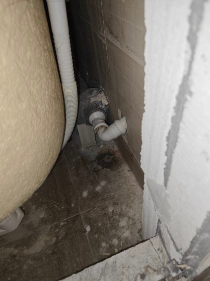 Biała rura odpływowa podłączona do wanny, widoczna przy szarej ścianie z płytkami i surowym wykończeniem. Ujęcie z dołu, blisko podłogi.