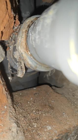 Szara rura kanalizacyjna z etykietą, zamontowana w murze z cegły. Widoczne ślady korozji na łączeniu rury. Ujęcie z bliska, w słabym oświetleniu.