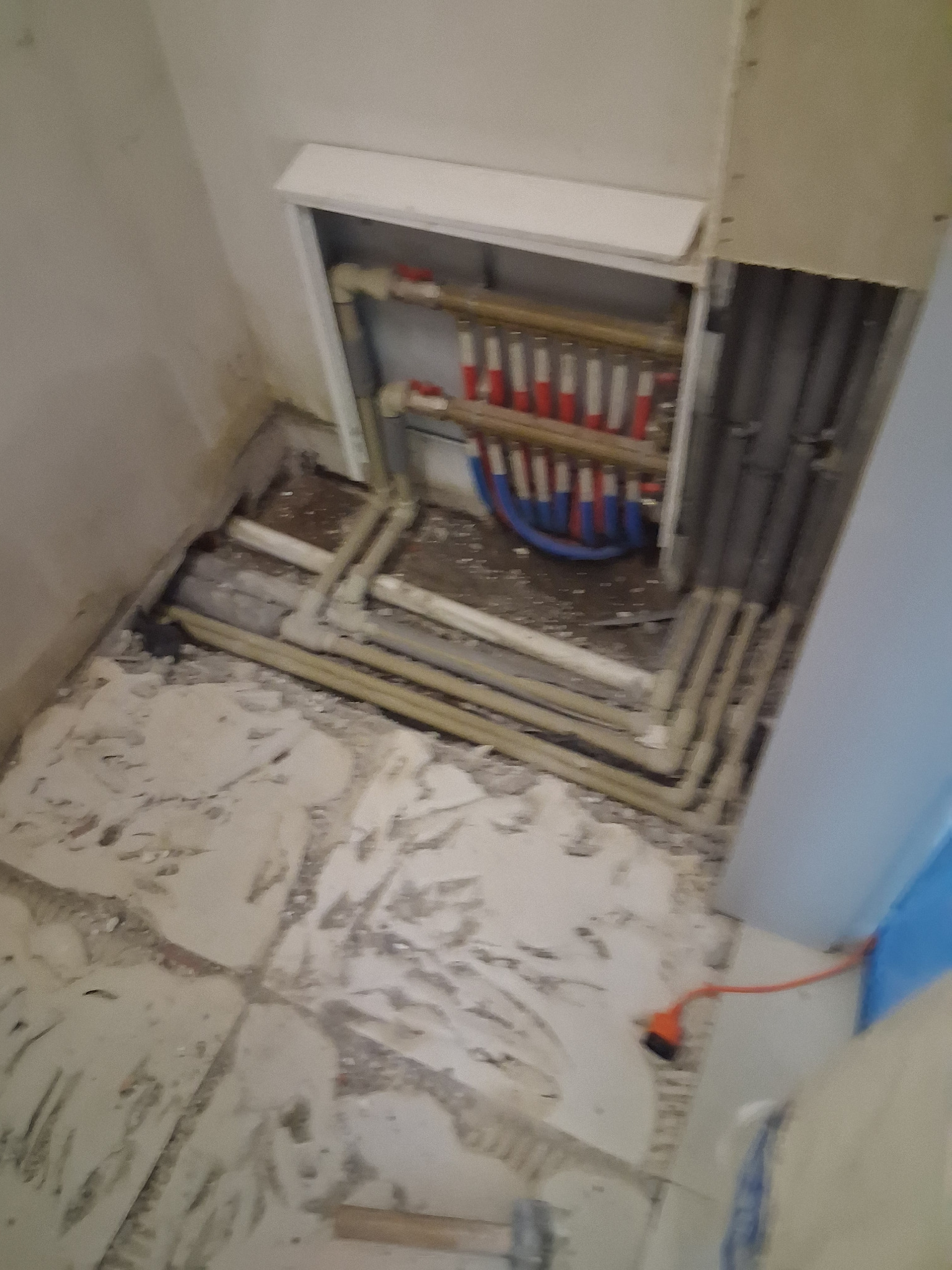 Rozdzielacz ogrzewania podłogowego w szafce, z widocznymi rurami doprowadzającymi i odprowadzającymi wodę. Podłoga w trakcie prac wykończeniowych, widoczne ślady po kleju.