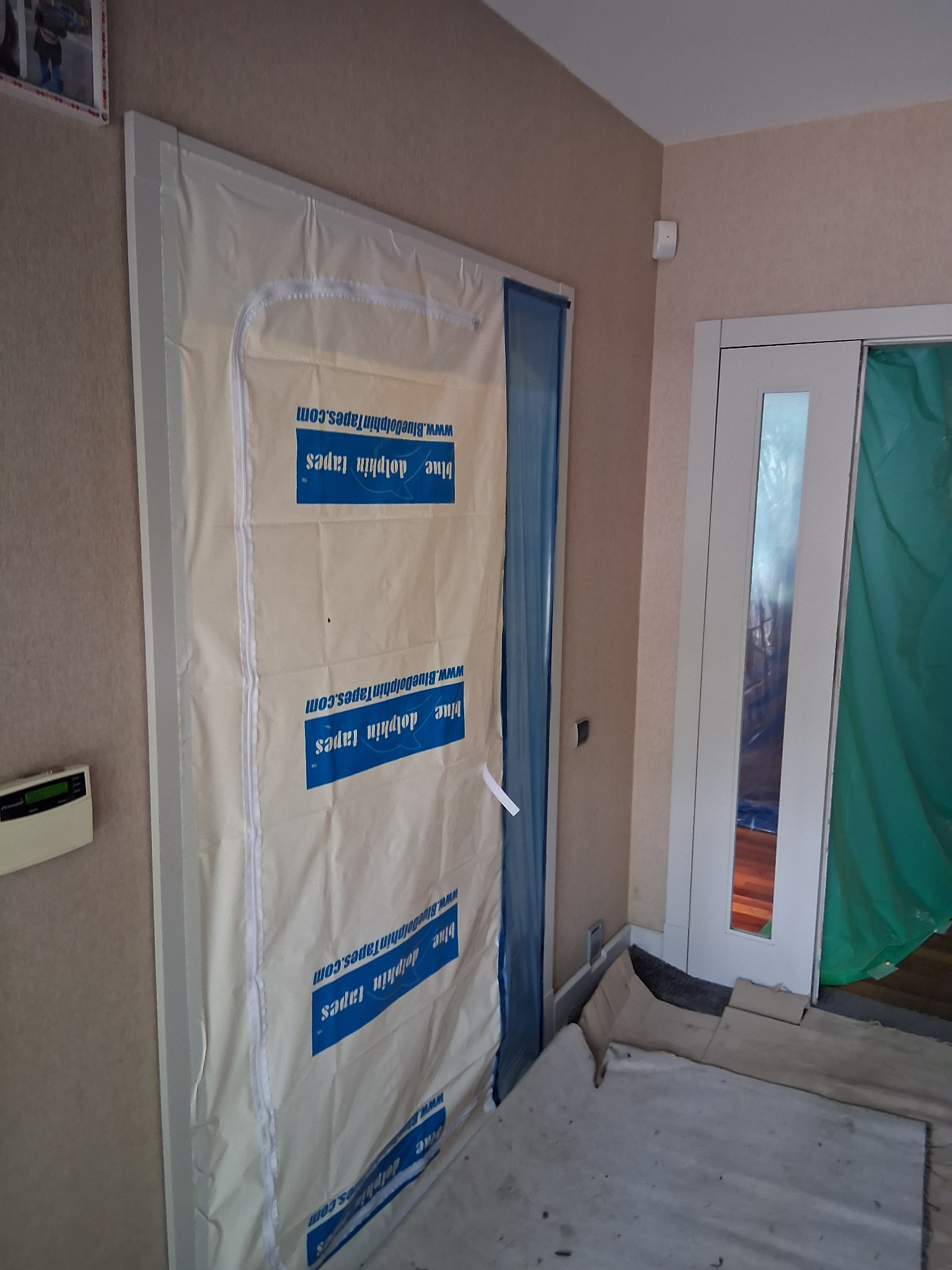 Zabezpieczone folią malarską drzwi z futryną podczas prac remontowych. Ochrona przed zabrudzeniem farbą. Widoczne taśmy Blue Dolphin. Przygotowanie do malowania.
