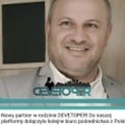 NIERUCHOMOŚCI-NOCLEGI SPÓŁKA Z OGRANICZONĄ ODPOWIEDZIALNOŚCIĄ - Uśmiechnięty mężczyzna w jasnej marynarce, z logo firmy deweloperskiej w tle, prezentowany jako nowy partner w branży pośrednictwa z Polską.